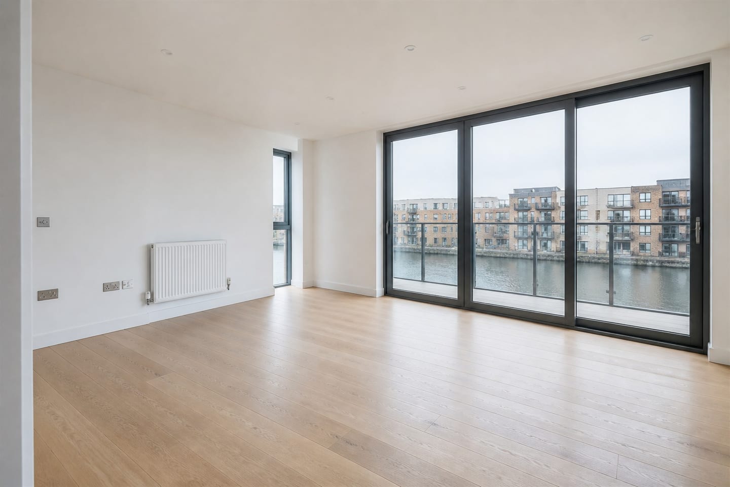 Empty waterfront new-build living room with floor-to-ceiling glazing onto a dock, shown before and after virtual staging with a tan leather sofa, steel-framed chairs and a reclaimed-wood coffee table — before