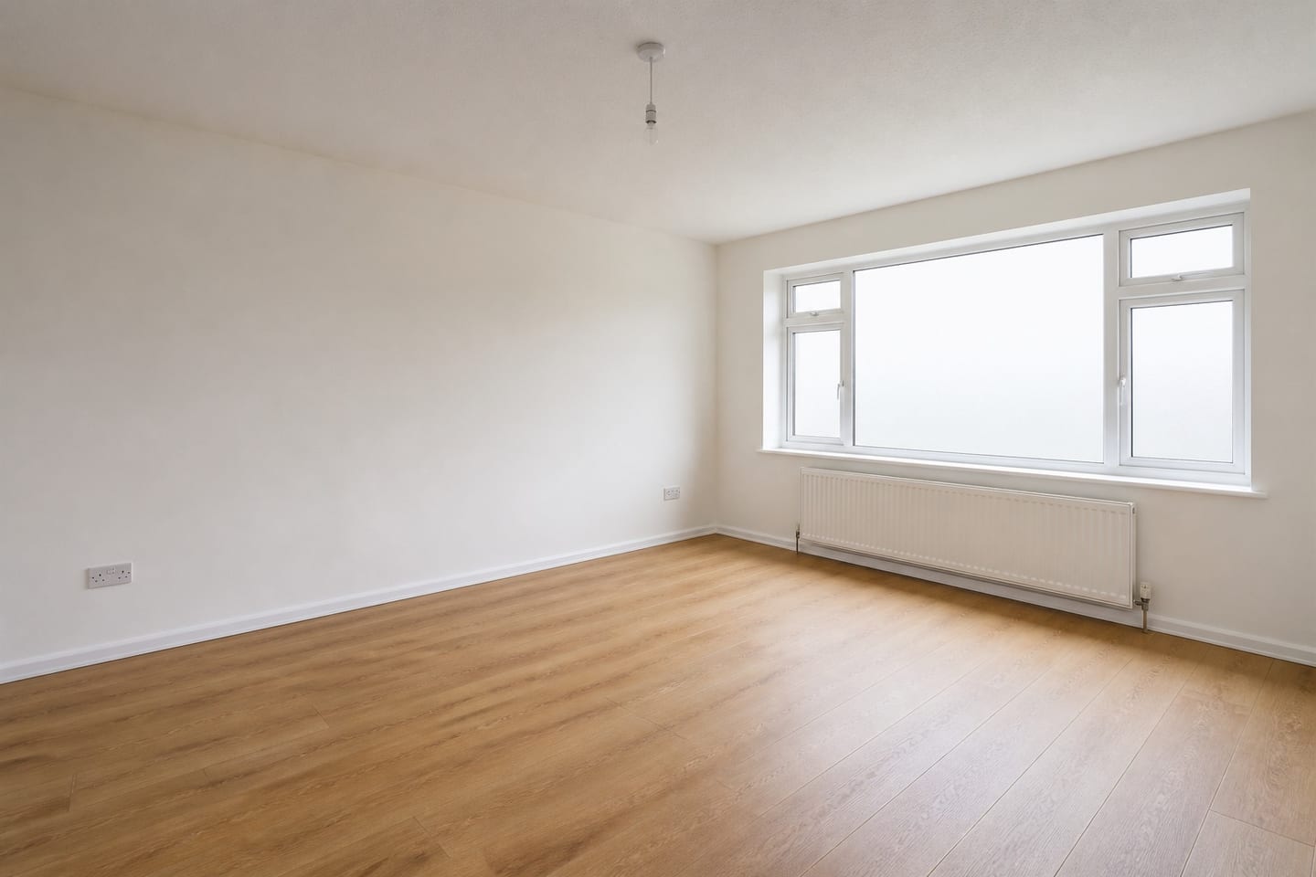 Empty rental living room with a wide casement window, radiator and oak-effect laminate floor, shown before and after virtual staging with a pale sofa, armchair, coffee table and framed prints — before