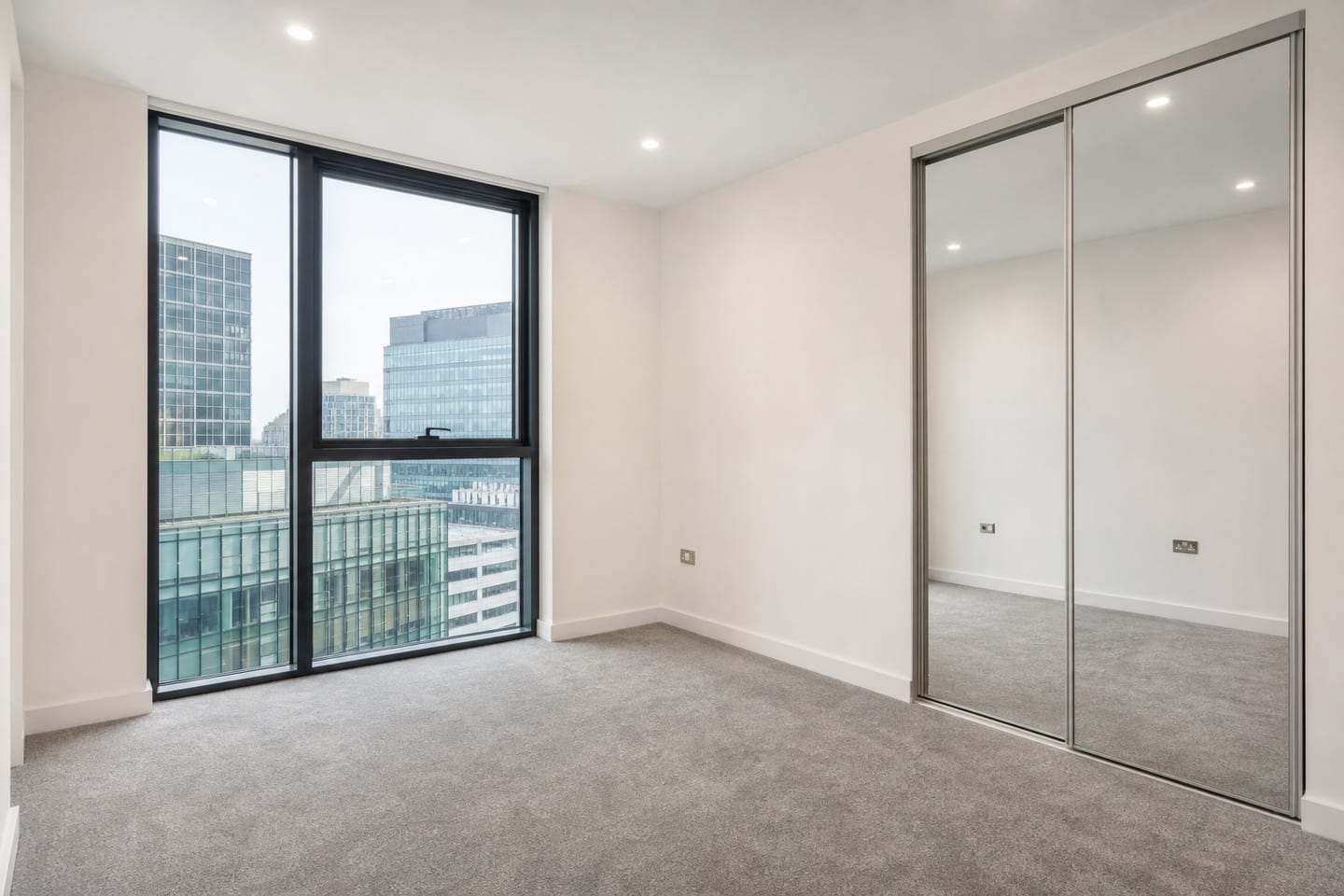 Empty compact London new-build double bedroom with full-height dark-framed glazing onto glass office blocks, a mirrored sliding wardrobe and grey carpet, shown before and after virtual staging with a low upholstered bed, oak nightstand, table lamp and jute rug — before