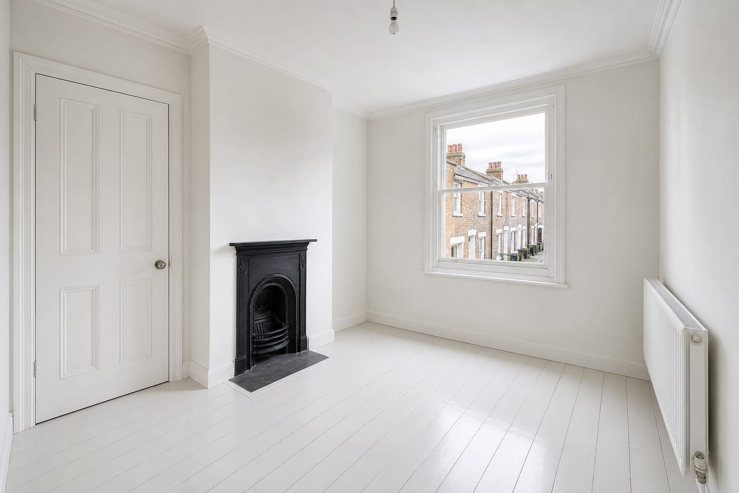 Empty compact London flat-conversion living room with a small cast-iron fireplace, a sash window onto stock-brick terraces and painted white floorboards, shown before and after virtual staging with a two-seater sofa, oak-framed armchair, small oval coffee table and jute rug — before
