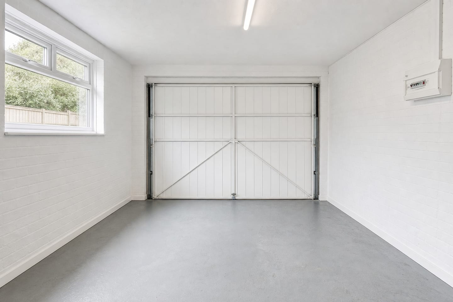 Empty converted garage with painted white blockwork, a boarded up-and-over door, a consumer unit and a bare concrete floor, shown before and after virtual staging with an exercise bike, weight bench, dumbbell rack and a leaning mirror — before