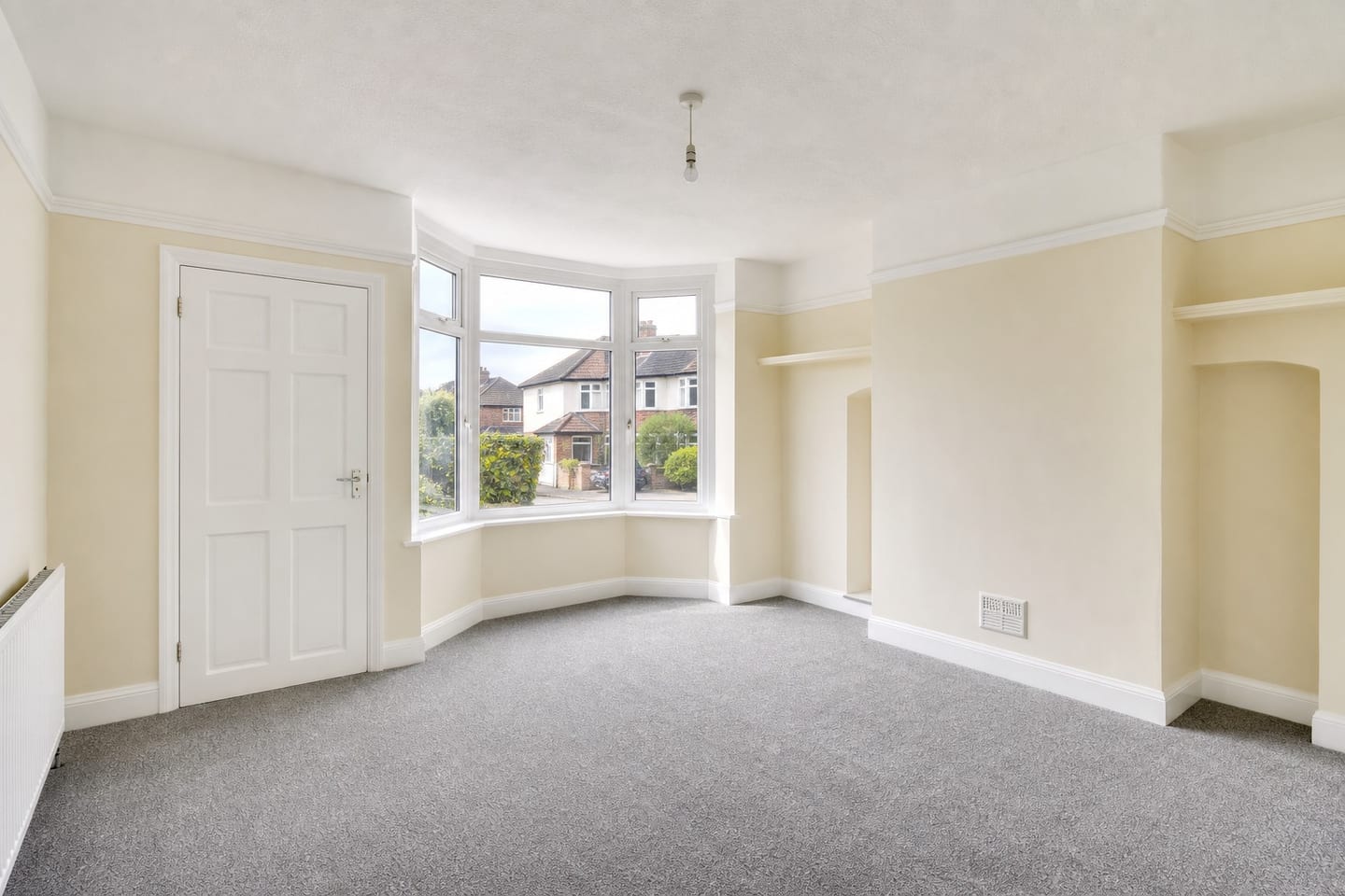 Empty 1930s semi-detached living room with a square bay window, chimney breast, alcove shelves and grey carpet, shown before and after virtual staging with a pale sofa, green armchair, walnut coffee table and side table — before