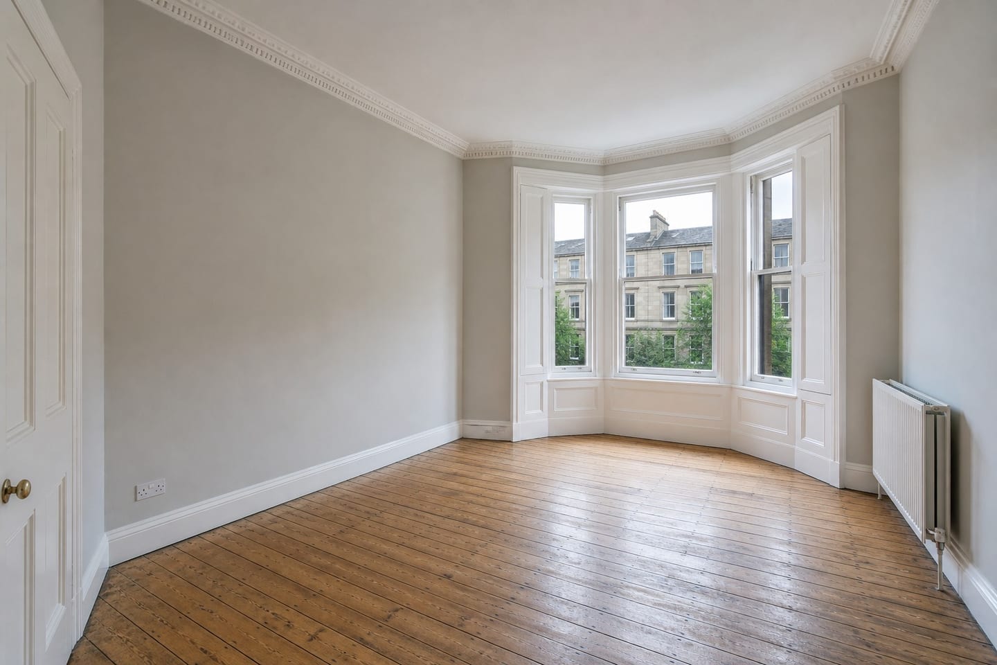 Empty Scottish tenement living room with a three-pane bay window, ornate cornicing and stripped floorboards, shown before and after virtual staging with a pale sofa, armchair and jute rug — before