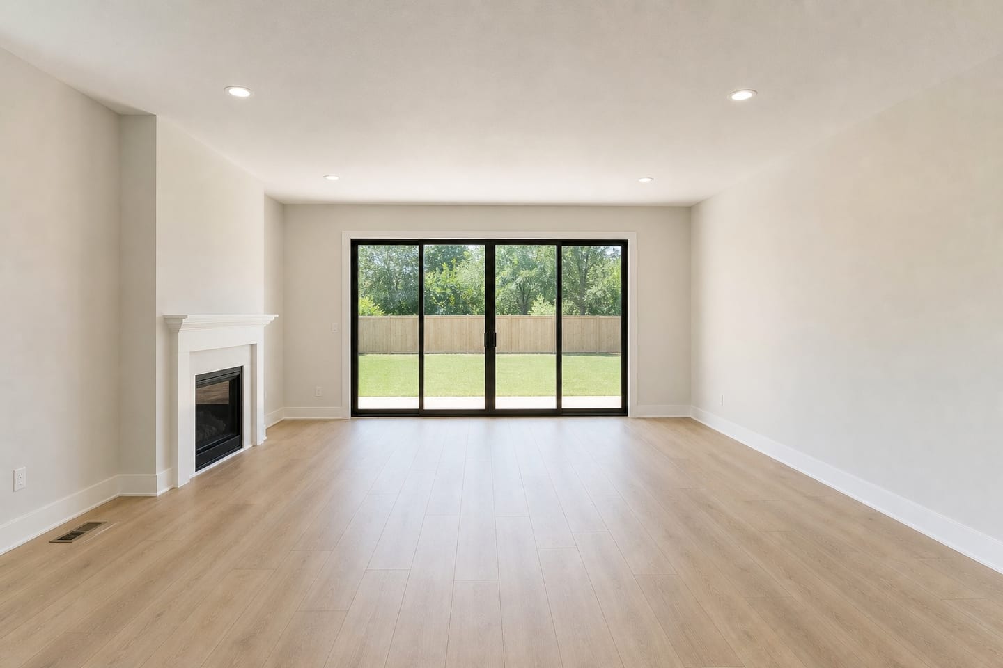 Empty American living room with a wide sliding glass patio door onto a fenced backyard, a white-mantel fireplace, recessed cans and wide-plank flooring, shown before and after virtual staging with a sofa, armchair, round coffee table, console and jute rug — before