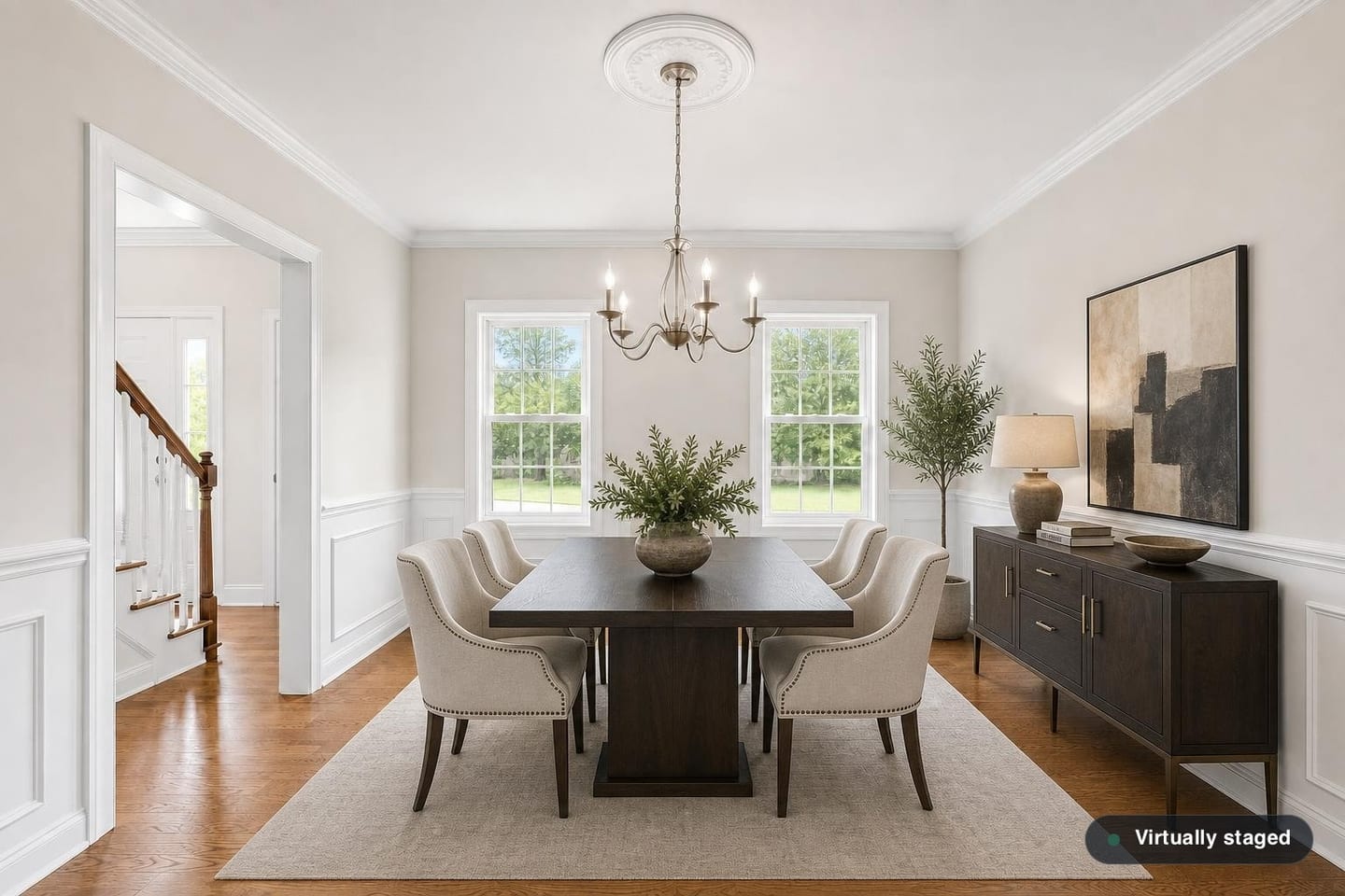 Empty formal dining room in a colonial house with wainscoting, a chair rail, crown molding, a five-arm chandelier on a ceiling medallion and a cased opening onto the staircase, shown before and after virtual staging with a rectangular table, six upholstered chairs, a sideboard and a rug — after