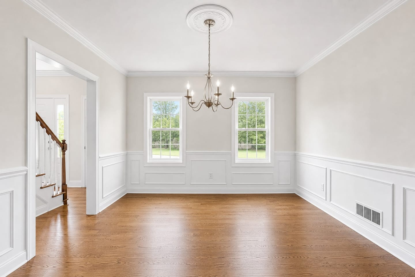 Empty formal dining room in a colonial house with wainscoting, a chair rail, crown molding, a five-arm chandelier on a ceiling medallion and a cased opening onto the staircase, shown before and after virtual staging with a rectangular table, six upholstered chairs, a sideboard and a rug — before