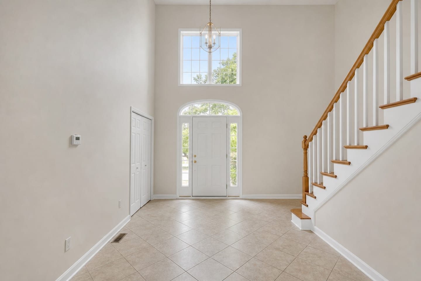 Empty two-story entry foyer with a front door flanked by sidelights under an arched fanlight, a hanging lantern fixture, a staircase with oak treads and white balusters, a bifold coat closet and diagonal tile flooring, shown before and after virtual staging with a console table, lamp, leaning mirror, coat stand, a bench with baskets and a runner — before