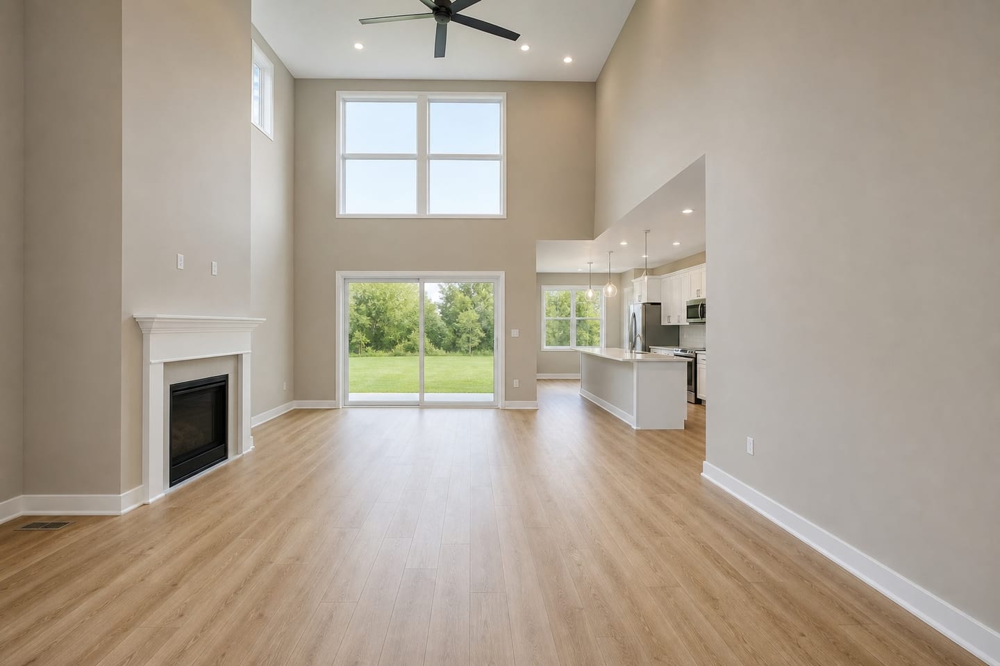 Empty two-story family room open to a kitchen, with a stacked clerestory window above a patio slider, a white-mantel fireplace, a ceiling fan and an empty island beyond the half-wall, shown before and after virtual staging with a sofa, armchair, coffee table and rug — before