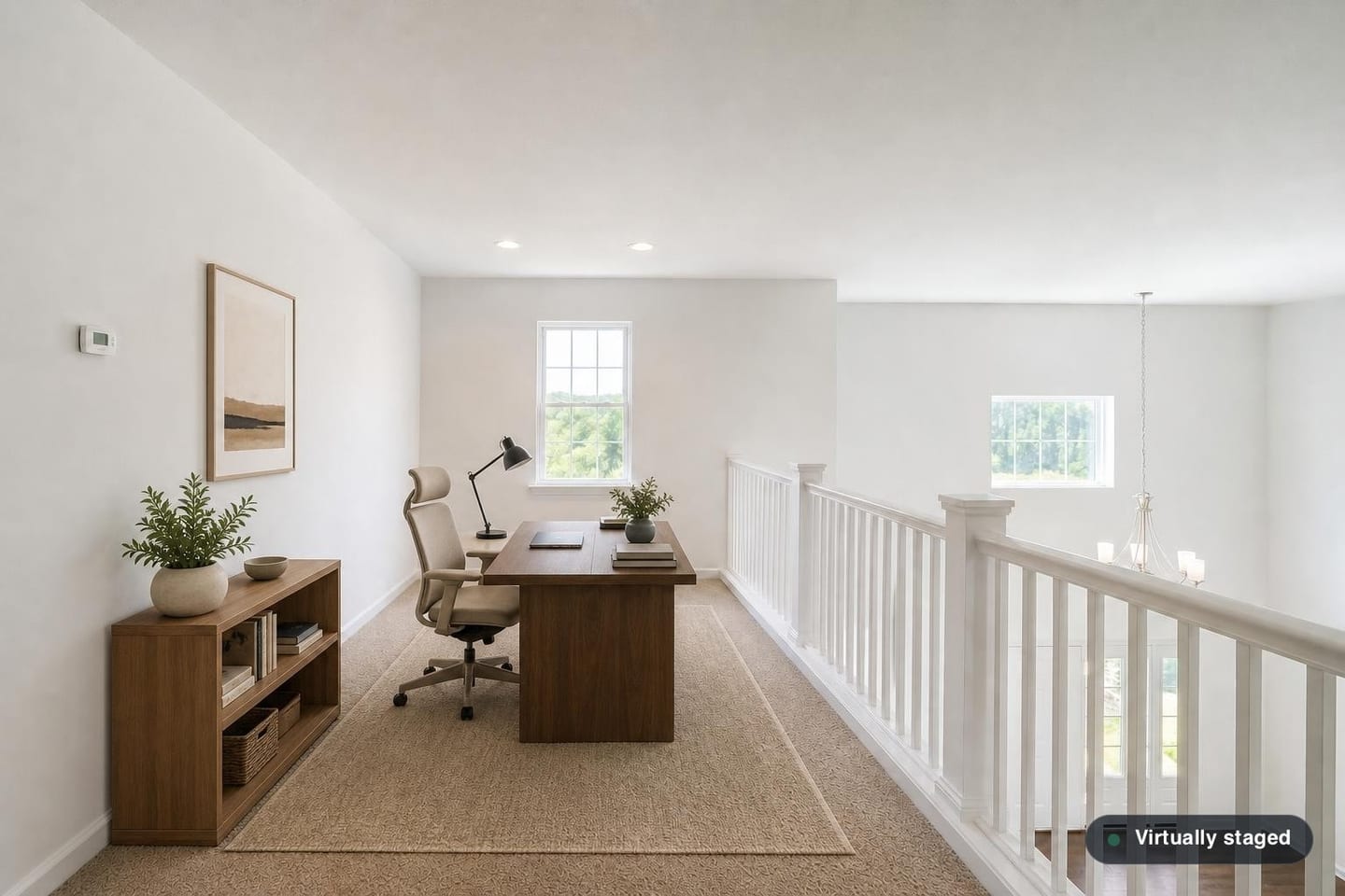 Empty open loft at the top of a staircase overlooking a two-story entry hall, with a white railing and square balusters, one double-hung window and beige carpet, shown before and after virtual staging with a desk, task chair, low bookcase and rug — after