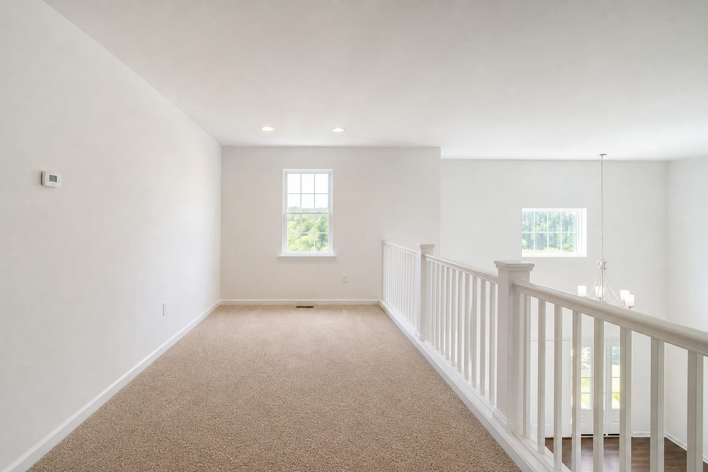 Empty open loft at the top of a staircase overlooking a two-story entry hall, with a white railing and square balusters, one double-hung window and beige carpet, shown before and after virtual staging with a desk, task chair, low bookcase and rug — before