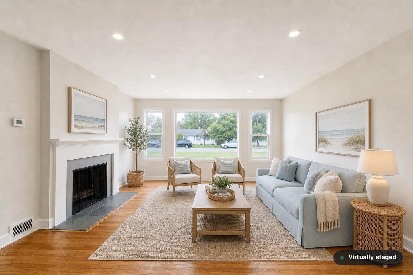 The same empty ranch front living room, shown before and after virtual staging in a Coastal scheme with a pale blue sofa, cane-backed chairs, a light oak coffee table, a woven side table and framed shore prints — after