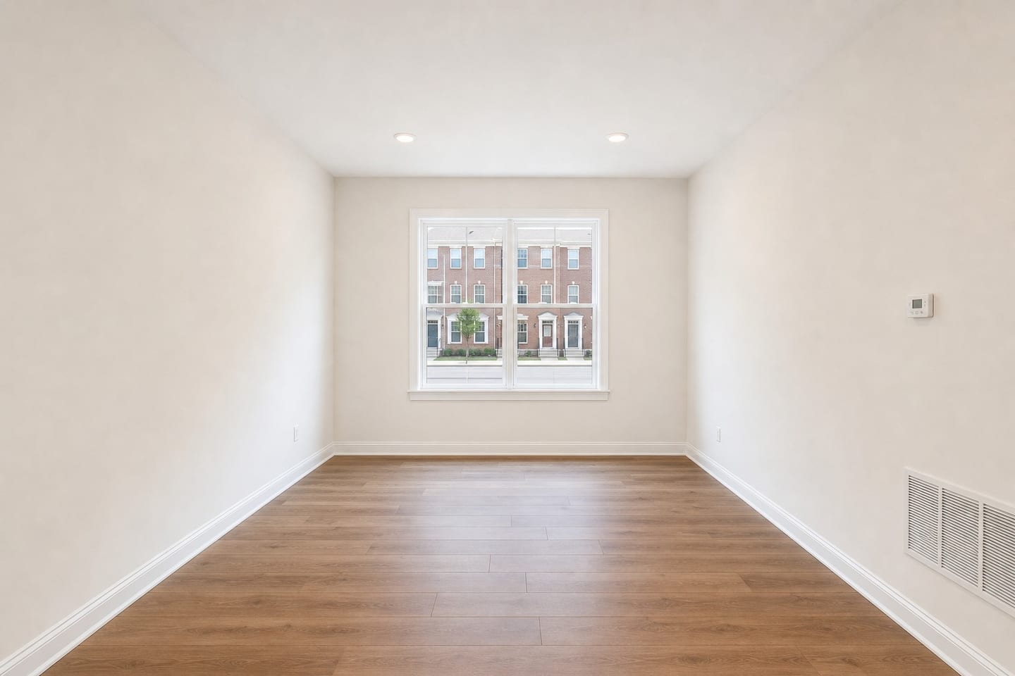 Empty narrow townhome living room with one window looking onto matching brick townhomes across the street, a wall return grille, a thermostat and engineered wood flooring, shown before and after virtual staging with a sofa, armchair, walnut coffee table and side table — before